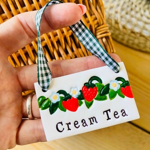 May include: White ceramic sign with a green gingham ribbon. The sign has a border of red and green strawberries and white flowers. The text on the sign reads "Cream Tea".