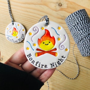 May include: Round white ceramic ornament featuring a cartoon bonfire design with a smiling face, stars, and decorative lines. The text "Bonfire Night" is written below. A black and white string is attached for hanging. The image also shows a roll of string.