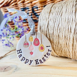 May include: White ceramic ornament with the text "Happy Easter!" and a design of colourful Easter eggs hanging from strings.