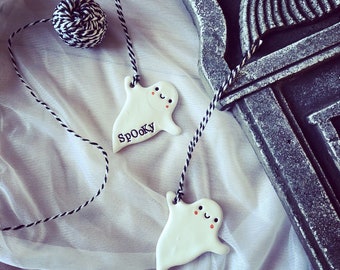 May include: Two white ceramic ghost ornaments with black and white twine strings. Each ghost features a smiling face with pink cheeks. One ghost has the word Spooky printed in black letters. The ornaments are attached to a ball of twine.