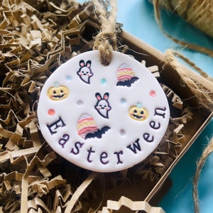 May include: White ceramic ornament with a brown twine hanger. The ornament is decorated with a variety of Halloween and Easter themed images, including pumpkins, ghosts, bats, and Easter eggs. The text "Easterween" is written in black lettering around the edge of the ornament.