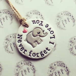 May include: A white ceramic ornament with a grey elephant and the text "I love you never forget" in black. A small pink heart is above the elephant.