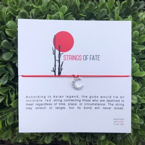 May include: Red string bracelet with a silver crescent moon charm. The bracelet is tied around a card with the text "STRINGS OF FATE" and a description of the legend of the red string of fate.