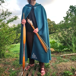 Op de afbeelding: Een persoon draagt een turquoise capuchon met witte borduursels en houdt een boog en pijlen vast. De cape heeft een decoratief ontwerp aan de onderkant.