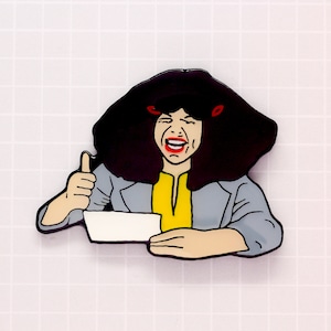 May include: A cartoon enamel pin of a woman with black hair and a gray jacket giving a thumbs up. She is holding a white rectangle in her hand and has a big smile on her face.