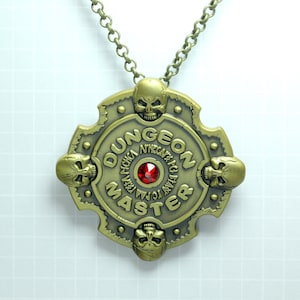 May include: Gold-toned metal necklace with a round pendant featuring the words "Dungeon Master" in a circular design with four skull accents and a red gemstone in the center.