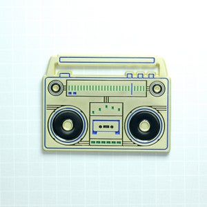 Puede incluir: Pin de metal dorado de un reproductor de casetes vintage con detalles azules y verdes. El reproductor de casetes tiene un diseño en blanco y negro con un dial y botones negros.
