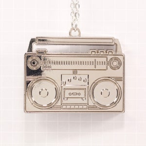 May include: A silver-toned boombox charm necklace. The charm is shaped like a vintage boombox with a cassette player, radio dial, and speakers.
