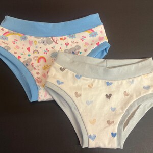Peut inclure: Deux culottes. L'une a un fond blanc avec un motif de koalas, d'arcs-en-ciel et de cœurs, bordée de bleu. L'autre a un fond blanc avec des cœurs bleus, marron et gris, bordée de bleu clair.