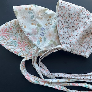 Peut inclure: Trois bonnets pour bébés à motifs floraux. Les bonnets présentent des imprimés floraux pastel en rose, vert et blanc. Chaque bonnet a une fermeture à nouer. Les bonnets sont en tissu doux.