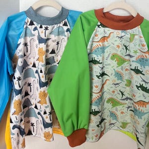 May include: Two long-sleeve art smocks with dinosaur patterns. The left smock has blue sleeves, a grey collar, and a white body with blue, yellow, and black dinosaurs. The right smock has green sleeves, a brown collar, and a light green dinosaur print.