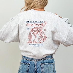 Puede incluir: Una sudadera gris con un estampado gráfico rojo y blanco de un vaquero montando un caballo salvaje. El gráfico incluye el texto "Daniel Ricciardo Honey Badger Never Left 19 89 High Performance Athlete".