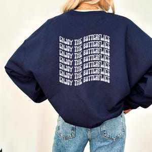 Può includere: Una felpa blu navy con il testo "ENJOY THE BUTTERFLIES" ripetuto in un font retrò sul retro.
