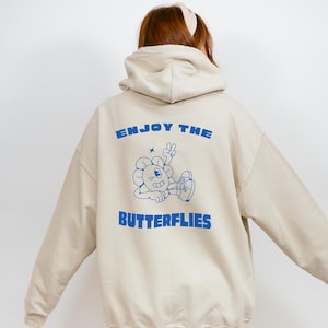 Può includere: Una felpa con cappuccio color crema con una grafica blu di un fiore dei cartoni animati con un viso e il testo "Enjoy the Butterflies" sul retro.