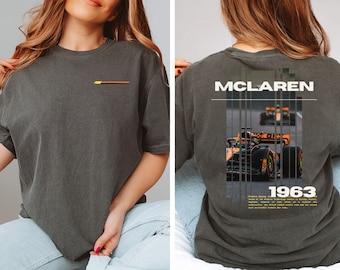May include: A dark grey t-shirt featuring a graphic of a McLaren race car and the text "McLaren 1963". The text below the graphic describes the car's history.