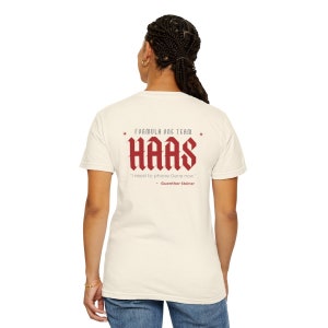 May include: A white t-shirt with a red graphic on the back. The graphic says "Formula One Team Haas" in a bold, gothic font. Below the name is a quote in a smaller font: "I need to phone Gene now." - Guenther Steiner.