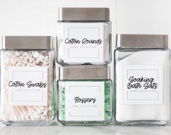 May include: Four glass jars with silver lids, labeled for home organization. The jars contain cotton swabs, cotton rounds, green flossers, and bath salts. Each jar has a white label with black text. The text on the labels reads "Cotton Swabs", "Cotton Rounds", "Hossers", and "Soaking Bath Salts".