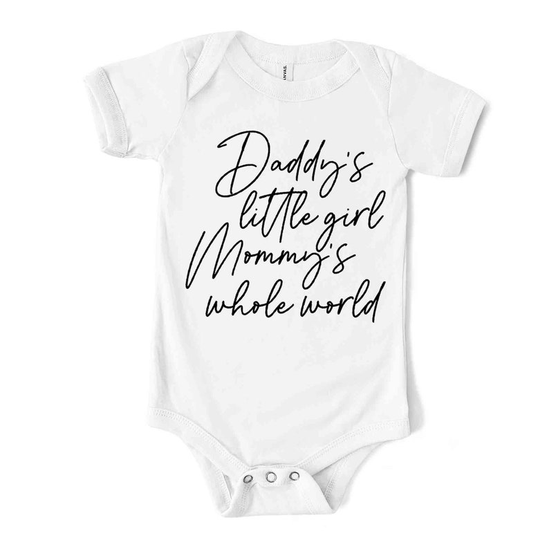 May include: White baby bodysuit with short sleeves. The text "Daddy's little girl Mommy's whole world" is written in black script across the front. The bodysuit has snap closures at the bottom.