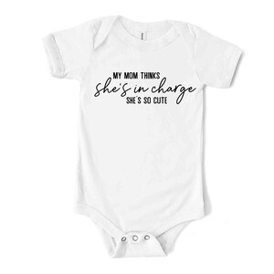 May include: White baby bodysuit with short sleeves. The text "MY MOM THINKS she's in charge SHE'S SO CUTE" is printed in black script across the front. The bodysuit has three poppers at the bottom.
