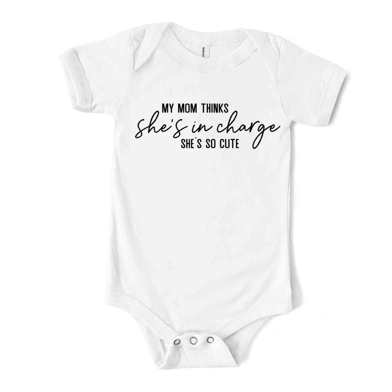 May include: White baby bodysuit with short sleeves. The text "MY MOM THINKS she's in charge SHE'S SO CUTE" is printed in black script across the front. The bodysuit has three poppers at the bottom.