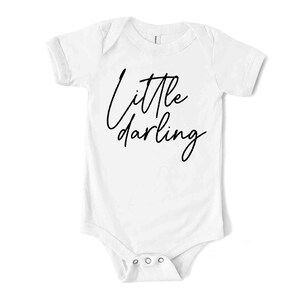May include: A white baby bodysuit with short sleeves and a snap closure. The words "Little darling" are written in black cursive script across the front. The bodysuit is designed for infants.