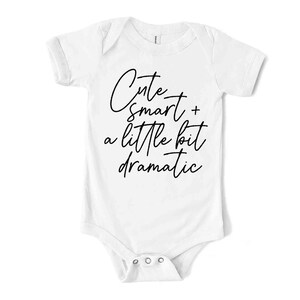 May include: White baby bodysuit with short sleeves and popper fastenings. The bodysuit has the text "Cute + smart + a little bit dramatic" in black script.