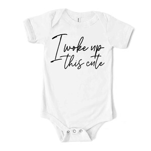 May include: White baby bodysuit with short sleeves. The bodysuit features the phrase "I woke up this cute" in black script. It has three poppers at the bottom for easy changing.
