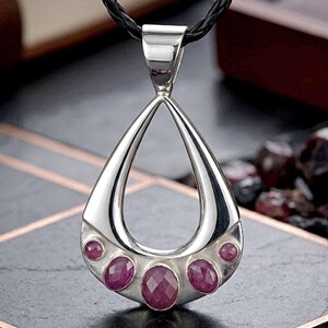 May include: A teardrop-shaped silver pendant featuring five bezel-set, oval-cut, deep pinkish-red gemstones along the bottom curve. The pendant is suspended from a black braided cord, showcasing a polished metallic finish and a symmetrical design.