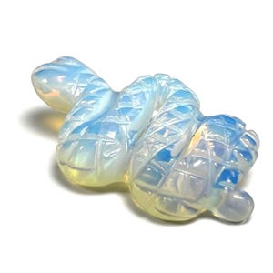 May include: A translucent, milky white opalite figurine carved into the shape of a coiled snake. The surface features a detailed, etched cross-hatch pattern across the body, with a smooth head and tail, exhibiting iridescent blue and yellow light reflections.