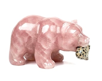 Puede incluir: Una figura de oso de cuarzo rosa pulido de pie sobre sus cuatro patas. El oso tiene una superficie rosa translúcida y lisa, y sostiene en su boca una pequeña concha marina de color tostado con manchas de color marrón oscuro.
