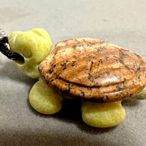May include: A carved green and brown turtle pendant with a black cord. The turtle is made of stone and has a detailed shell.