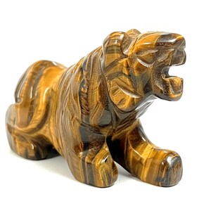 May include: A carved tiger figurine made of tiger's eye stone. The sculpture features a detailed depiction of a tiger in a crouching position with its mouth open. The stone has brown and gold hues.