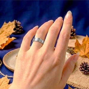Op de afbeelding: Een zilveren ring met een geometrisch ontwerp en een kleine, heldere steen wordt aan een vinger gedragen. De ring staat tegen een achtergrond van herfstbladeren en een donkerblauwe ondergrond.