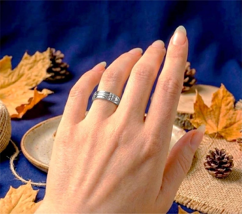 Op de afbeelding: Een zilveren ring met een geometrisch ontwerp en een kleine, heldere steen wordt aan een vinger gedragen. De ring staat tegen een achtergrond van herfstbladeren en een donkerblauwe ondergrond.