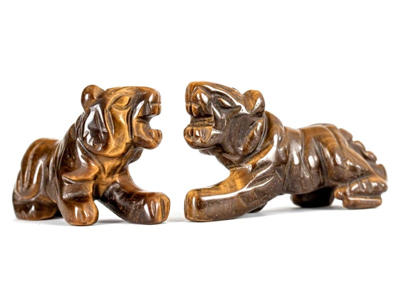May include: Two carved tiger figurines in shades of brown and amber are displayed against a white background. The tigers are posed in a roaring stance, showcasing detailed craftsmanship. These decorative sculptures are ideal for home decor.
