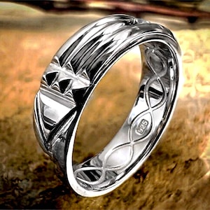 Op de afbeelding: Een zilveren ring met een gedetailleerd ontwerp. De ring heeft geometrische patronen en een getextureerd oppervlak. De binnenkant van de ring is gestempeld met "925". De ring wordt gepresenteerd op een bruin oppervlak.