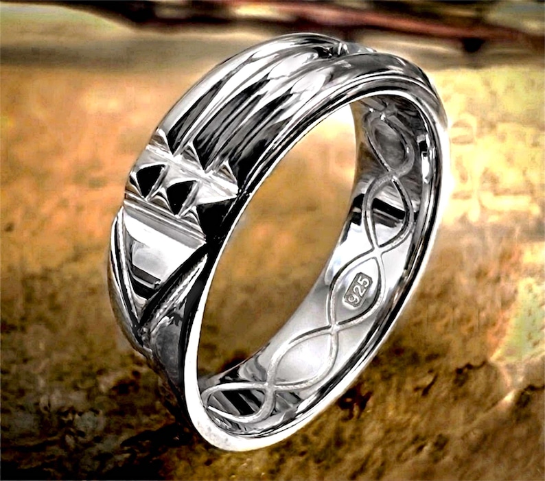Op de afbeelding: Een zilveren ring met een gedetailleerd ontwerp. De ring heeft geometrische patronen en een getextureerd oppervlak. De binnenkant van de ring is gestempeld met "925". De ring wordt gepresenteerd op een bruin oppervlak.