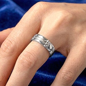 Op de afbeelding: Een zilveren ring met geometrische ontwerpen wordt aan een vinger gedragen. De ring heeft een reeks ribbels en driehoekige patronen. De achtergrond is een donkerblauwe stof.