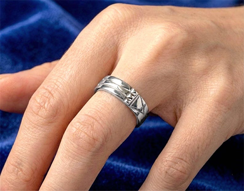 Op de afbeelding: Een zilveren ring met geometrische ontwerpen wordt aan een vinger gedragen. De ring heeft een reeks ribbels en driehoekige patronen. De achtergrond is een donkerblauwe stof.