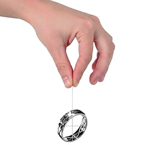 Op de afbeelding: Een zilveren ring met een geometrisch ontwerp hangt aan een dunne draad. De ring heeft ingewikkelde patronen en een gepolijste afwerking, die licht reflecteert. Het ontwerp van de ring omvat stervormige vormen en gebogen lijnen.