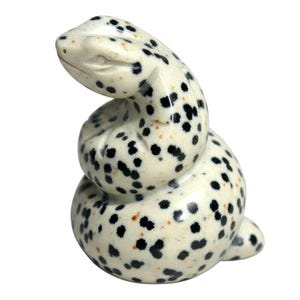 Könnte beinhalten: Eine kleine, zusammengerollte Schlangenfigur aus cremefarbenem Stein mit einem gesprenkelten Muster aus schwarzen Punkten und dezenten orangefarbenen Flecken. Die Skulptur hat eine glatte, polierte Oberfläche mit geschnitzten Details.
