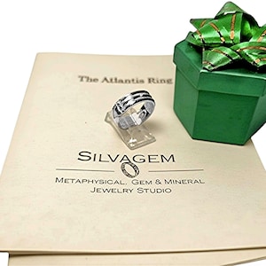 Op de afbeelding: Een zilveren ring met zwarte accenten wordt getoond op een transparante standaard, naast een groene geschenkdoos met een groene strik. De ring ligt op cr&egrave;mekleurig papier met de tekst "The Atlantis Ring" en "SILVAGEM". De sieradenstudio is "Metaphysical, Gem & Mineral".