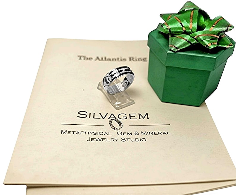 Op de afbeelding: Een zilveren ring met zwarte accenten wordt getoond op een transparante standaard, naast een groene geschenkdoos met een groene strik. De ring ligt op cr&egrave;mekleurig papier met de tekst "The Atlantis Ring" en "SILVAGEM". De sieradenstudio is "Metaphysical, Gem & Mineral".