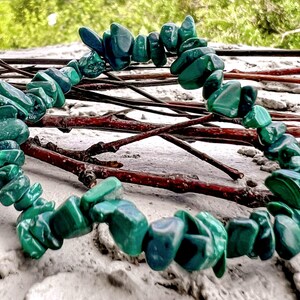 Op de afbeelding: Een armband gemaakt van gepolijste, onregelmatig gevormde groene malachietstenen. De stenen zijn aan elkaar geregen en vormen een cirkelvorm. De achtergrond omvat takjes en een zanderig oppervlak, met een vage groene achtergrond.