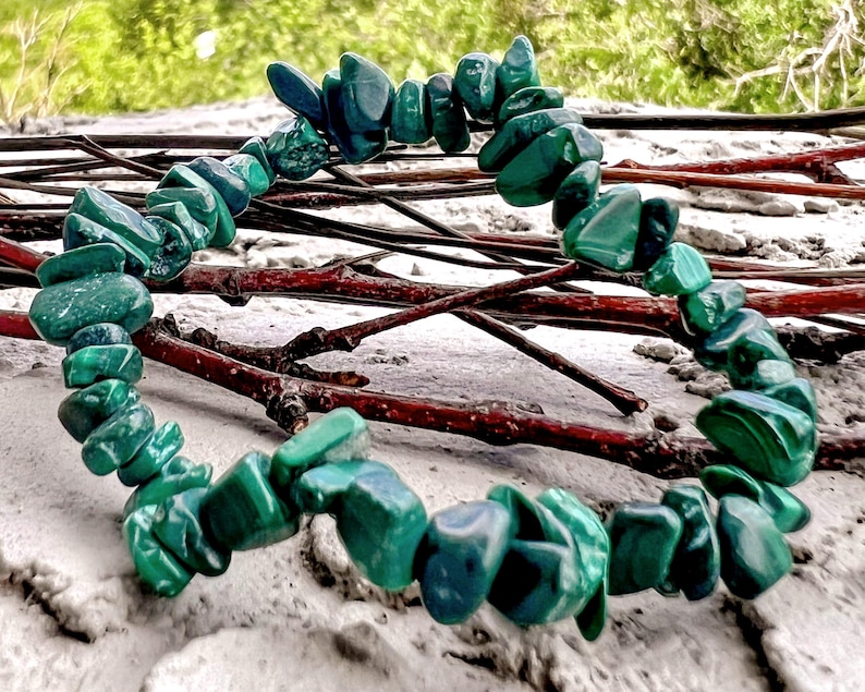 Op de afbeelding: Een armband gemaakt van gepolijste, onregelmatig gevormde groene malachietstenen. De stenen zijn aan elkaar geregen en vormen een cirkelvorm. De achtergrond omvat takjes en een zanderig oppervlak, met een vage groene achtergrond.