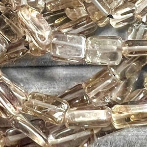 May include: Close-up of a strand of rectangular, translucent beads. The beads have a light brown hue and are strung together, creating a necklace or bracelet. The beads are evenly shaped and have a polished appearance.