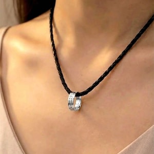 Op de afbeelding: Een zilveren ringhangerketting met een zwarte gevlochten koord. De ringhanger heeft een textuurpatroon. De ketting wordt om de nek gedragen.