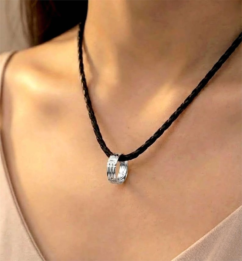 Op de afbeelding: Een zilveren ringhangerketting met een zwarte gevlochten koord. De ringhanger heeft een textuurpatroon. De ketting wordt om de nek gedragen.