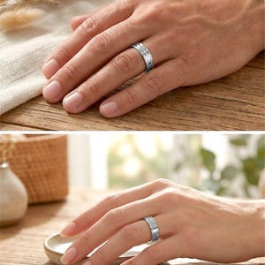 Op de afbeelding: Zilverkleurige ring met een hartvormig ontwerp en een getextureerde band. De ring wordt op twee handen getoond, wat suggereert dat het een partnerring is. De ring wordt gepresenteerd op een houten oppervlak.