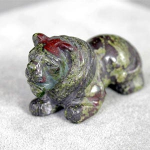 May include: A small, hand-carved stone figurine of a lion in a resting position. The sculpture features a mottled pattern of deep forest green, olive, and dark reddish-brown colors throughout the polished surface, highlighting the feline features.